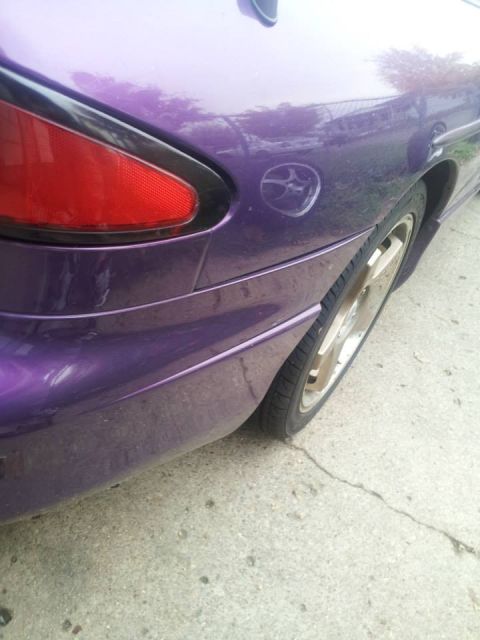 1995 Ford Probe - The rear quarter panel of a purple car is shown, featuring a red taillight and a circular emblem on the body. The vehicle is parked on a co…