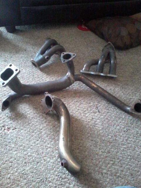 1995 Ford Probe - Two exhaust manifold headers are laid out on a carpeted floor. One is a polished metal piece with a curved shape, and the other is a darker…
