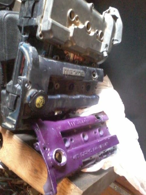 1995 Ford Probe - Three cast-aluminum cam covers/cylinder heads stacked on a wooden bench, two bearing 'Mazda' script and the bottom one painted purple readi…