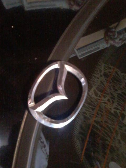 1995 Ford Probe - A close-up shot shows a silver Mazda emblem on a dark, reflective surface, possibly a car hood. The emblem is oval-shaped with a stylized w…