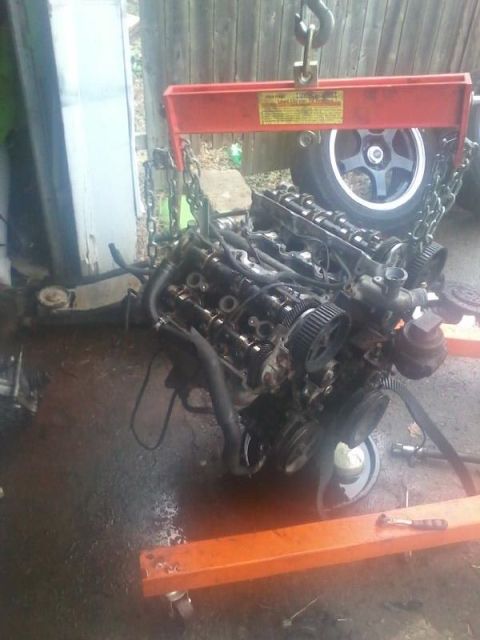 1995 Ford Probe - A V6 engine is suspended by a red engine hoist with chains. A car wheel is visible behind the engine, and the engine is resting on an orang…