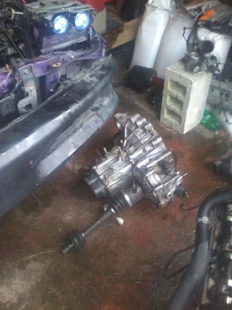 1995 Ford Probe - A disassembled vehicle transmission and drive axle are laid out on a wet, stained concrete floor. In the background, parts of a vehicle's f…