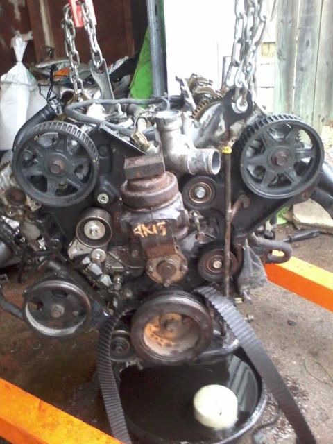 1995 Ford Probe - A disassembled engine, likely from a 1995 Ford Probe, is suspended by chains over an orange engine hoist. The engine features a timing belt…