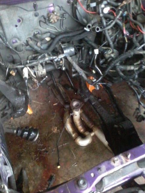 1995 Ford Probe - A close-up view of a disassembled car engine bay with various hoses, wires, and an exhaust manifold visible on the floor. The engine bay is…