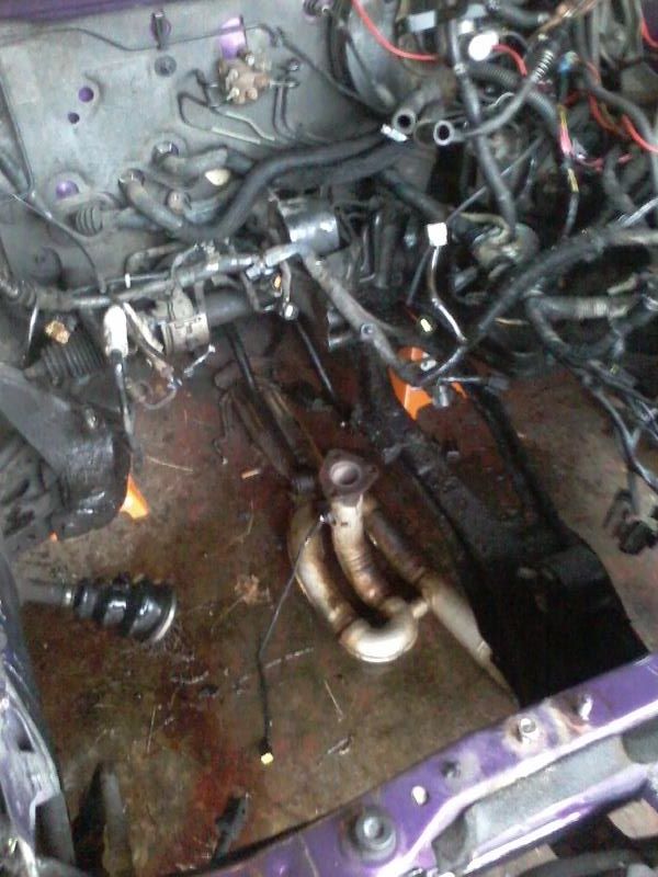 Disassembled car engine bay with hoses, wires, and exhaust manifold on the floor.