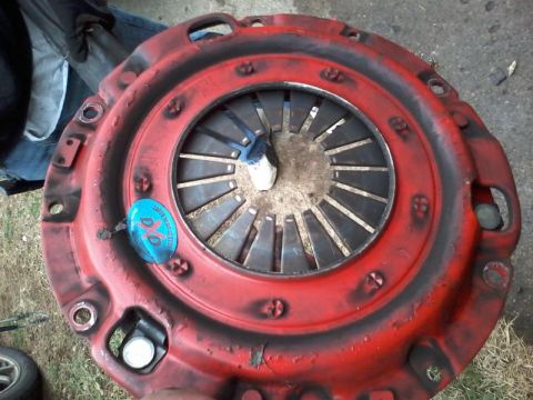 1995 Ford Probe - A red clutch pressure plate is shown, with a blue and white sticker on it. The part is resting on the ground.