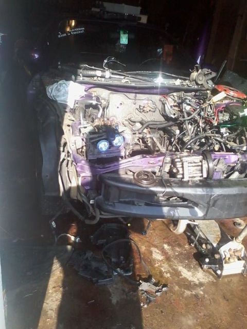 1995 Ford Probe - The front end of a purple car is disassembled, revealing the engine bay and headlights. Various car parts are scattered on the wet, concret…