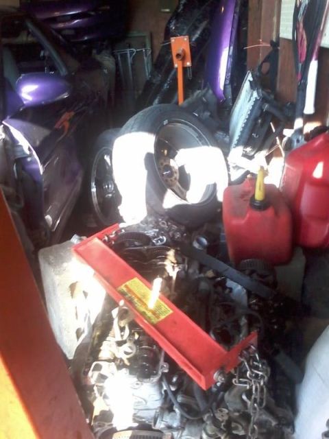 1995 Ford Probe - A disassembled car engine is in the foreground, with a red engine hoist positioned over it. In the background, parts of a purple car, inclu…