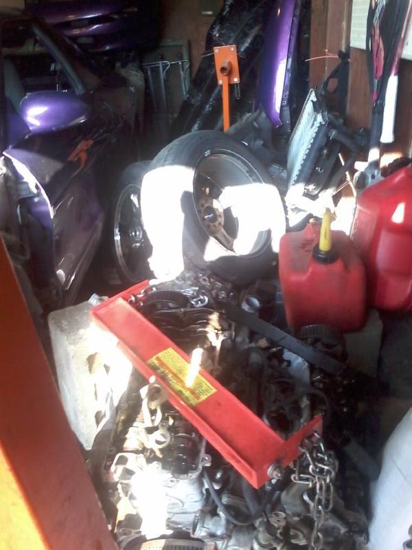 Disassembled car engine with red hoist, purple car parts, and gas cans in a cluttered garage.