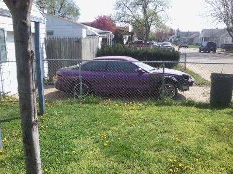 1995 Ford Probe - A purple coupe is parked in a grassy yard behind a chain-link fence. The front end of the car is damaged and missing parts.