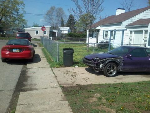 1995 Ford Probe - A damaged purple Ford Probe GT is parked on a driveway next to a white house. A red coupe is parked on the street in front of a chain-link…