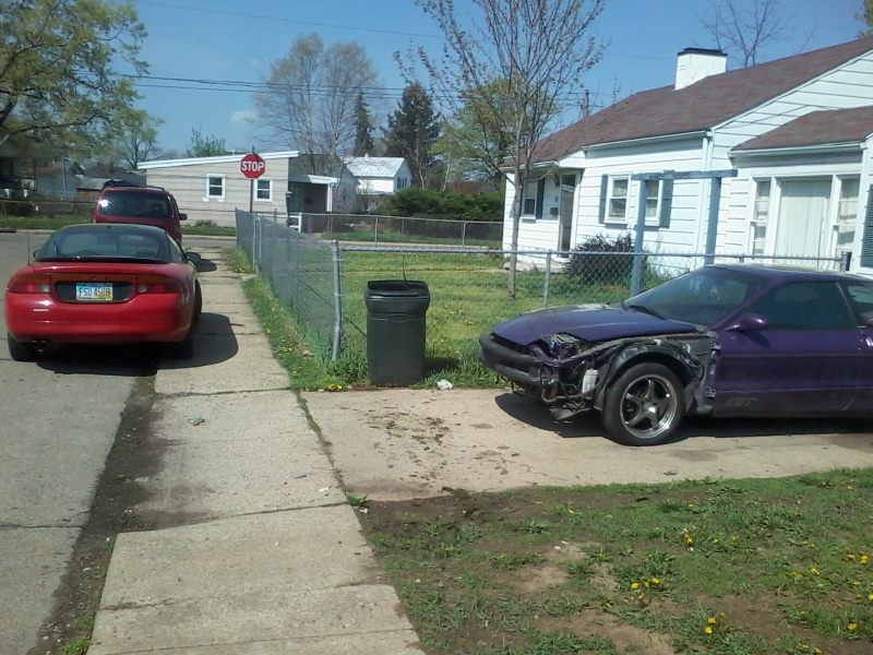 Damaged purple Ford Probe GT parked on driveway, red coupe on street.