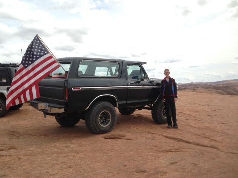 1979 Ford Bronco - A person stands next to a lifted black 1979 Ford Bronco with an American flag mounted on the rear. The vehicle is parked on a rocky, desert…