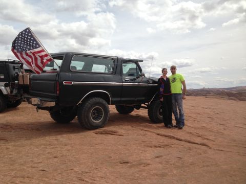 1979 Ford Bronco - A couple poses in front of a black 1979 Ford Bronco with an American flag flying from the tailgate. The Bronco is parked on a rocky, desert…
