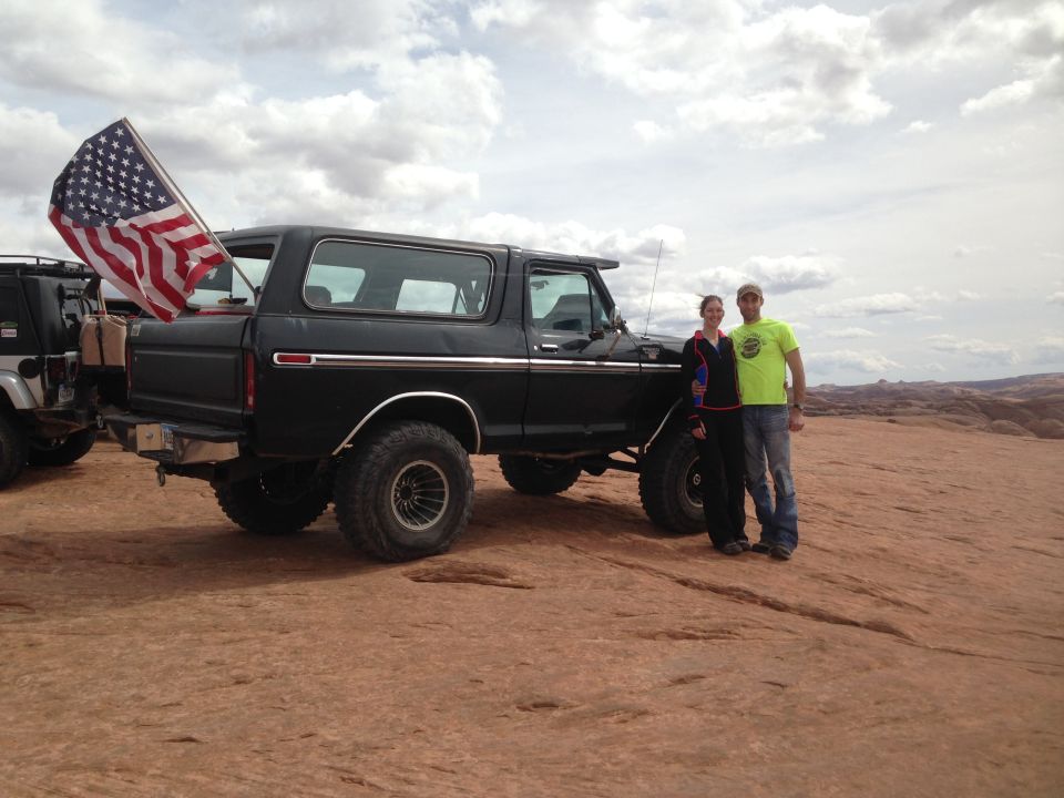 Couple posing in front of a black 1979 Ford Bronco with an American flag in a desert landscape.