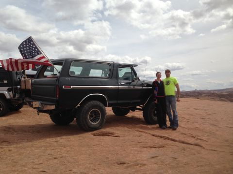 1979 Ford Bronco - A black 1979 Ford Bronco is parked on a rocky, desert landscape with an American flag mounted on the rear. A couple stands beside the vehic…