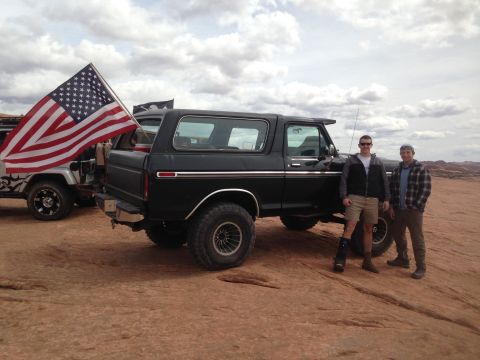 1979 Ford Bronco - Two men stand next to a black Ford Bronco with an American flag attached to a nearby Jeep. The Bronco is parked on a rocky, desert landscap…