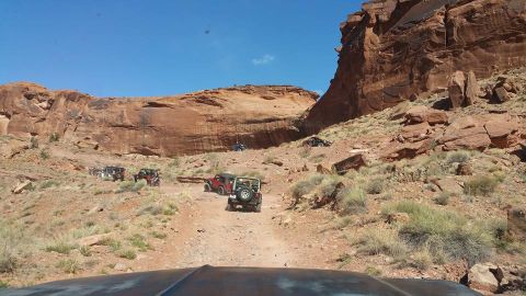 1979 Ford Bronco - A group of off-road vehicles, including several Jeep Wranglers, navigate a rocky, desert trail with large red rock formations in the backgr…
