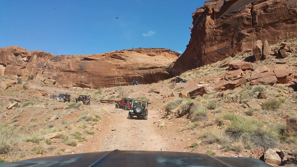 Off-road Jeeps driving on a rocky desert trail with red rock cliffs.