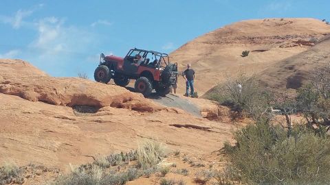 1979 Ford Bronco - A red Jeep Wrangler is driving up a steep, rocky incline in a desert landscape. Two people are standing near the Jeep, observing its progre…