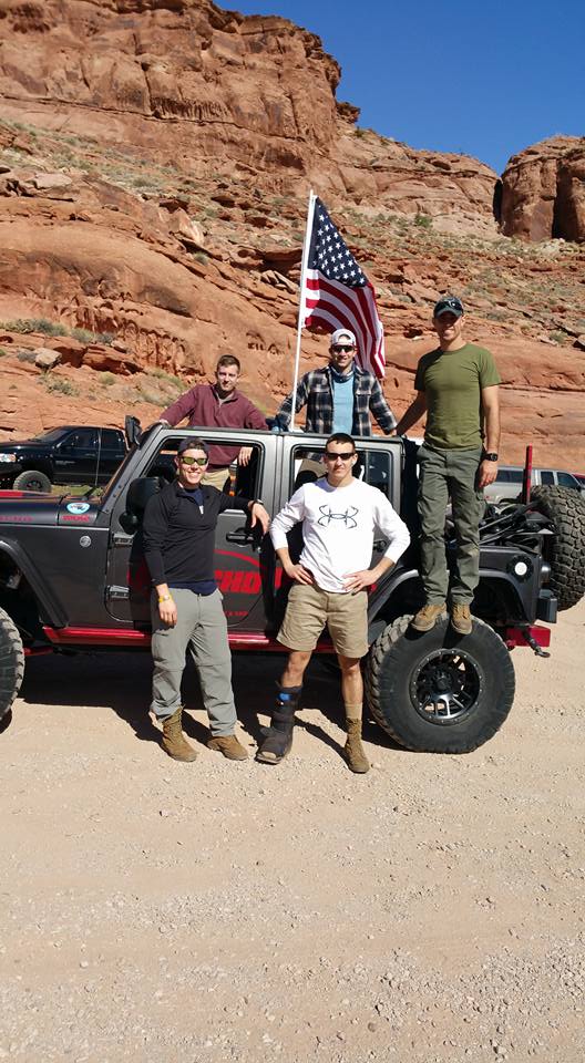 Five men posing with an American flag in front of a dark gray Jeep Wrangler in a desert landscape.