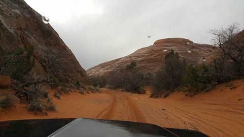 1979 Ford Bronco - The front hood of a vehicle is visible in the foreground, driving on a sandy, off-road trail through a desert landscape with red rock forma…