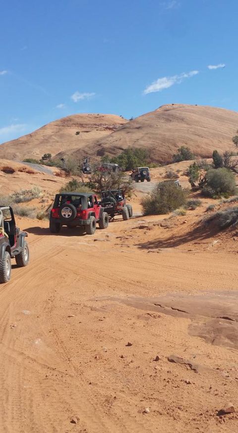 1979 Ford Bronco - A group of off-road vehicles, including several red Jeeps, navigate a sandy, desert trail with large rock formations and sparse vegetation…