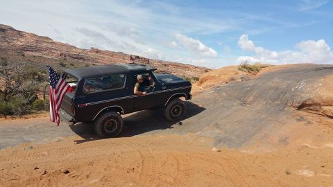 1979 Ford Bronco - A black, lifted 1970s Ford Bronco with an American flag attached to the rear is navigating a rocky, desert terrain under a partly cloudy sk…