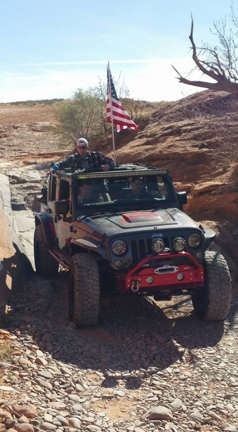 1979 Ford Bronco - A black Jeep Wrangler with an American flag mounted on the roof rack drives off-road on a rocky trail. A man wearing a baseball cap and sun…