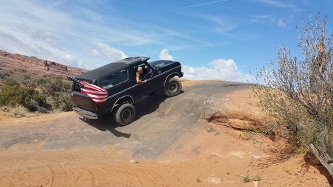 1979 Ford Bronco - A black Ford Bronco with an American flag in the tailgate is driving up a rocky incline in a desert landscape.