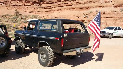1979 Ford Bronco - A black Ford Bronco with large off-road tires is parked on a dirt road in a desert landscape. An American flag is attached to the rear of t…