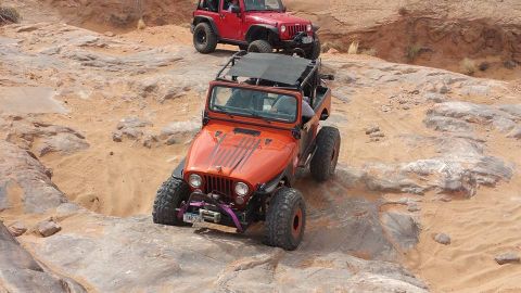 1979 Ford Bronco - Two off-road modified Jeeps navigate a rocky, sandy trail. The front Jeep is orange with black racing stripes on the hood and large off-roa…