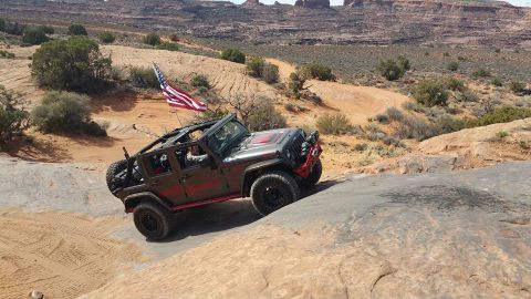 1979 Ford Bronco - A dark brown Jeep Wrangler with an American flag mounted on the back is navigating a rocky, off-road trail in a desert landscape.