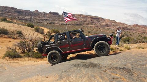 1979 Ford Bronco - A dark gray Jeep Wrangler with an American flag is driving off-road on a rocky incline. A man stands by the side of the trail, looking at h…