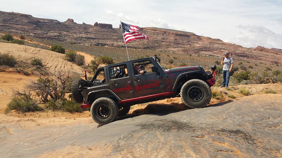 Dark gray Jeep Wrangler with American flag driving off-road on a rocky incline. Man standing by trail.
