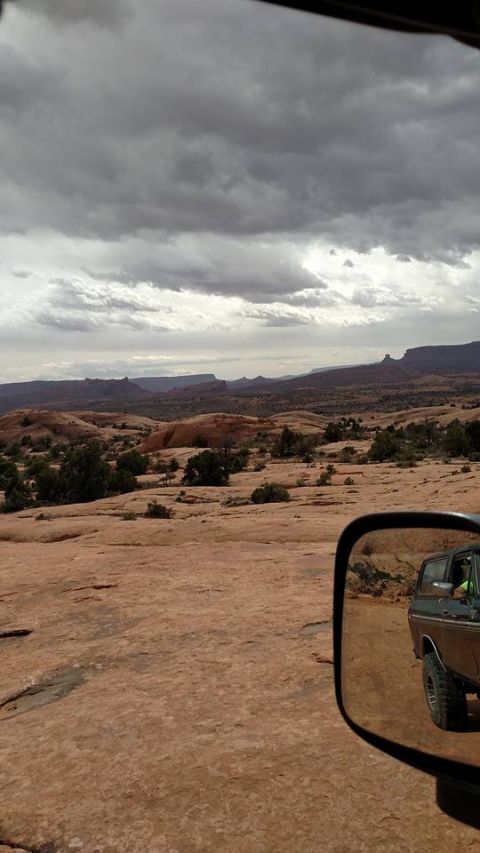 1979 Ford Bronco - The side view mirror of a dark colored SUV, likely a 1979 Ford Bronco, reflects the vehicle and the surrounding desert landscape. The vehic…