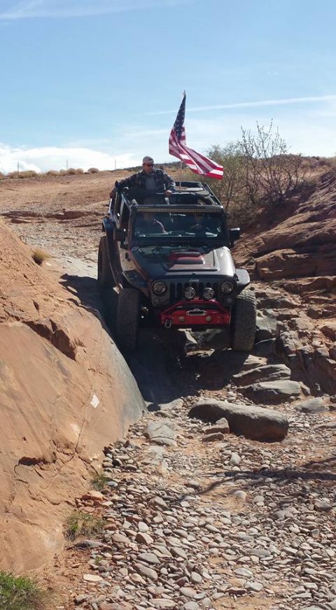 1979 Ford Bronco - A black Jeep Wrangler with an American flag flying from the roof rack navigates a rocky, off-road trail next to a large sandstone formation.