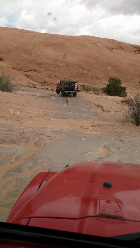 1979 Ford Bronco - A dark-colored SUV with an American flag attached to the rear is driving up a rocky, desert terrain. The front of a red vehicle is visible…
