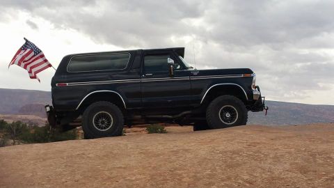 1979 Ford Bronco - A black, lifted 1979 Ford Bronco is parked on a rocky overlook with an American flag attached to the rear. The vehicle is viewed from the s…