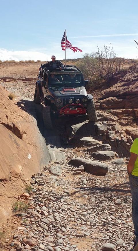 1979 Ford Bronco - A black Jeep Wrangler is navigating a rocky, off-road trail with an American flag flying from the roof. The driver is standing up through t…