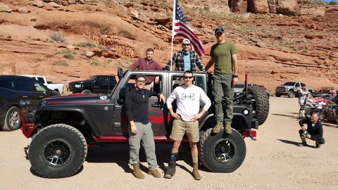 1979 Ford Bronco - A group of six men pose with a modified dark gray Jeep Wrangler in a desert landscape with red rock formations in the background. An Americ…