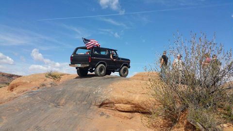 1979 Ford Bronco - A black 1979 Ford Bronco is parked on a rocky, desert landscape with an American flag flying from the rear. Several people are standing beh…