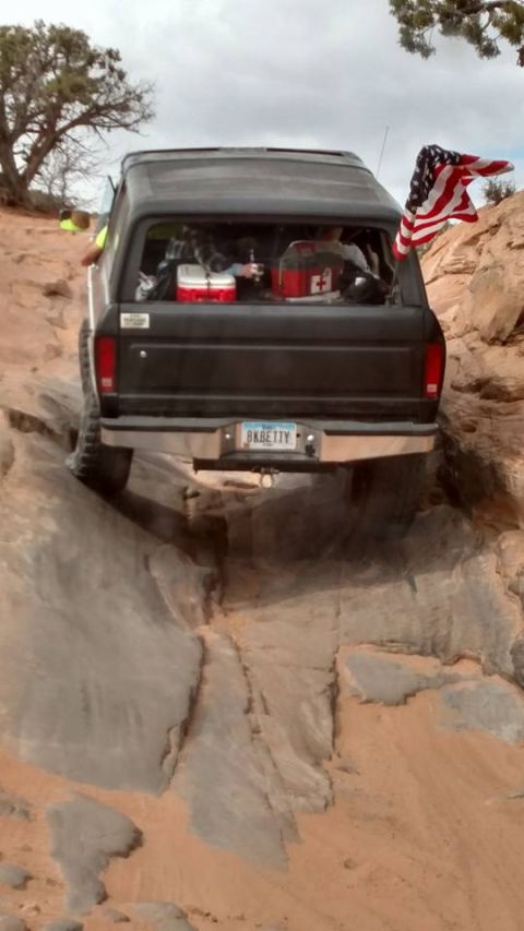 1979 Ford Bronco - The rear of a black 1979 Ford Bronco is shown navigating a rocky, off-road trail. An American flag is attached to the rear passenger side o…