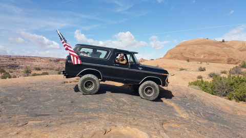 1979 Ford Bronco - A black Ford Bronco is driving on a rocky, desert landscape with an American flag attached to the rear. A man wearing a hat is visible driv…