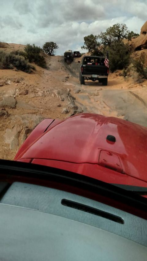 1979 Ford Bronco - The front of a red vehicle is visible in the foreground, looking out at a line of off-road vehicles driving up a rocky, dusty trail under a…