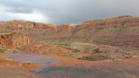 1979 Ford Bronco - A wide shot of a desert landscape with layered rock formations under a cloudy sky. The foreground features reddish-brown and dark gray rock…