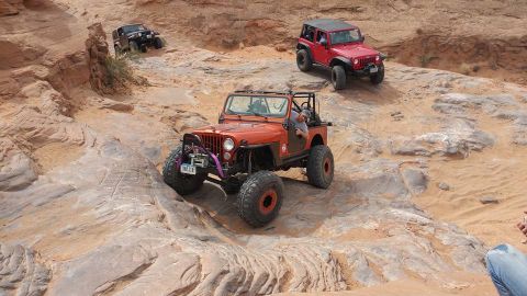 1979 Ford Bronco - An orange Jeep Wrangler is navigating a rocky, off-road trail, with a red Jeep Wrangler visible in the background. The orange Jeep has larg…