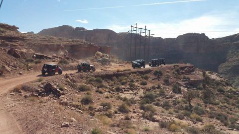 1979 Ford Bronco - A group of off-road vehicles, including a red Jeep Wrangler and several dark-colored SUVs, navigate a dusty dirt road through a rocky, dese…