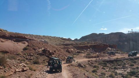 1979 Ford Bronco - Several off-road vehicles, including Jeeps, drive along a dusty dirt path through a rocky, desert landscape with red rock formations and a…