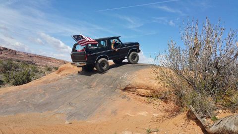 1979 Ford Bronco - A black 1979 Ford Bronco with an American flag displayed in the rear is driving on a rocky, desert terrain under a blue sky with scattered…